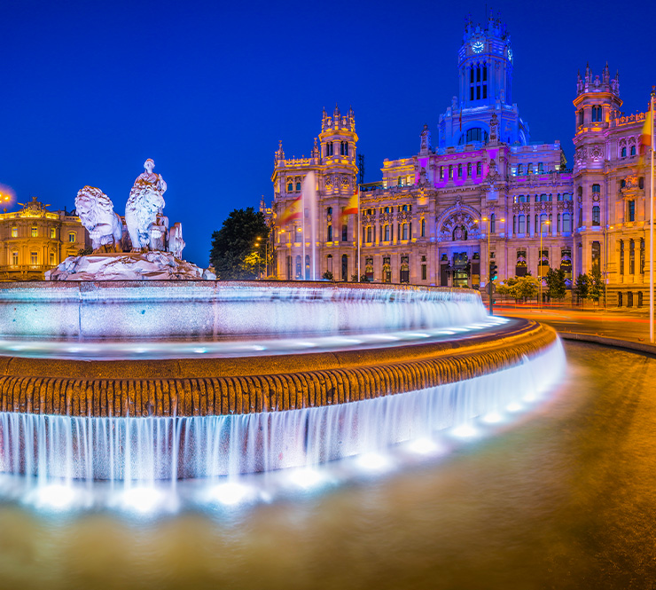 Spotlights illuminating the 18th Century fountain of Cybele overlooked by the iconic towers of the Palacio de Cibeles colourfully lit against the deep blue dusk skies above the Plaza de Cibele.