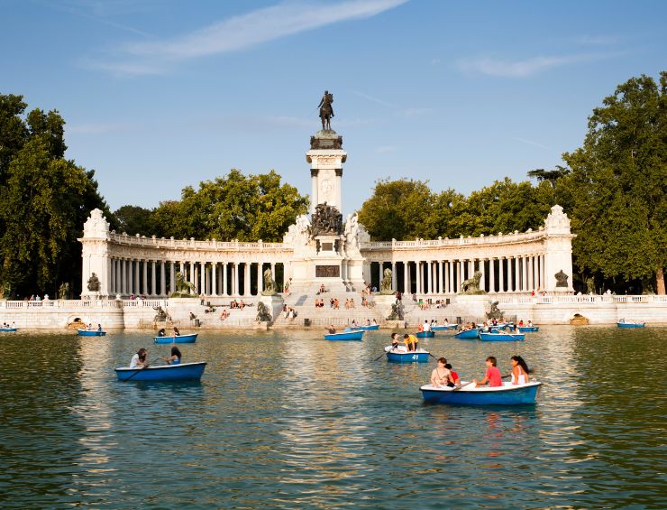 Groups of people row on blue boats across the Retiro pond in front of the Monument to Alfonso XI