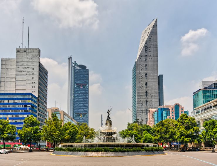 A statued fountain springs up in the middle of a city landscape with skyscrapers behind it.