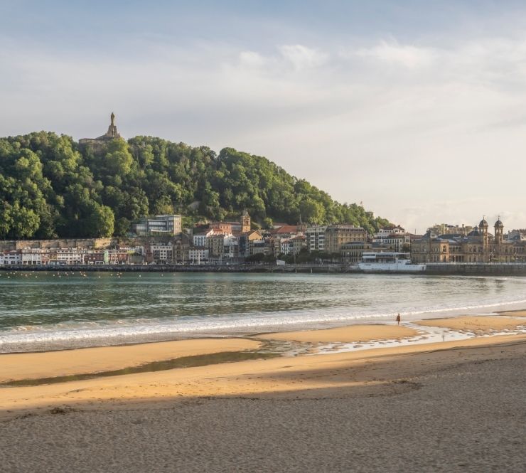 Old Spanish city buildings sit on a shoreline below a large tree-covered hill