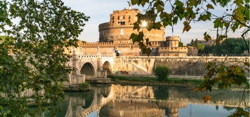 A stone castle at sunset reflecting onto a river, trees in the foreground