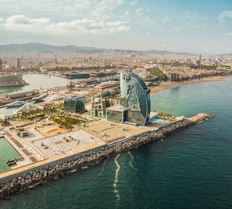 Aerial view of a half-circular skyscraper and other buildings on dock, shoreline and water in the background on a sunny day in Barcelona, Spain