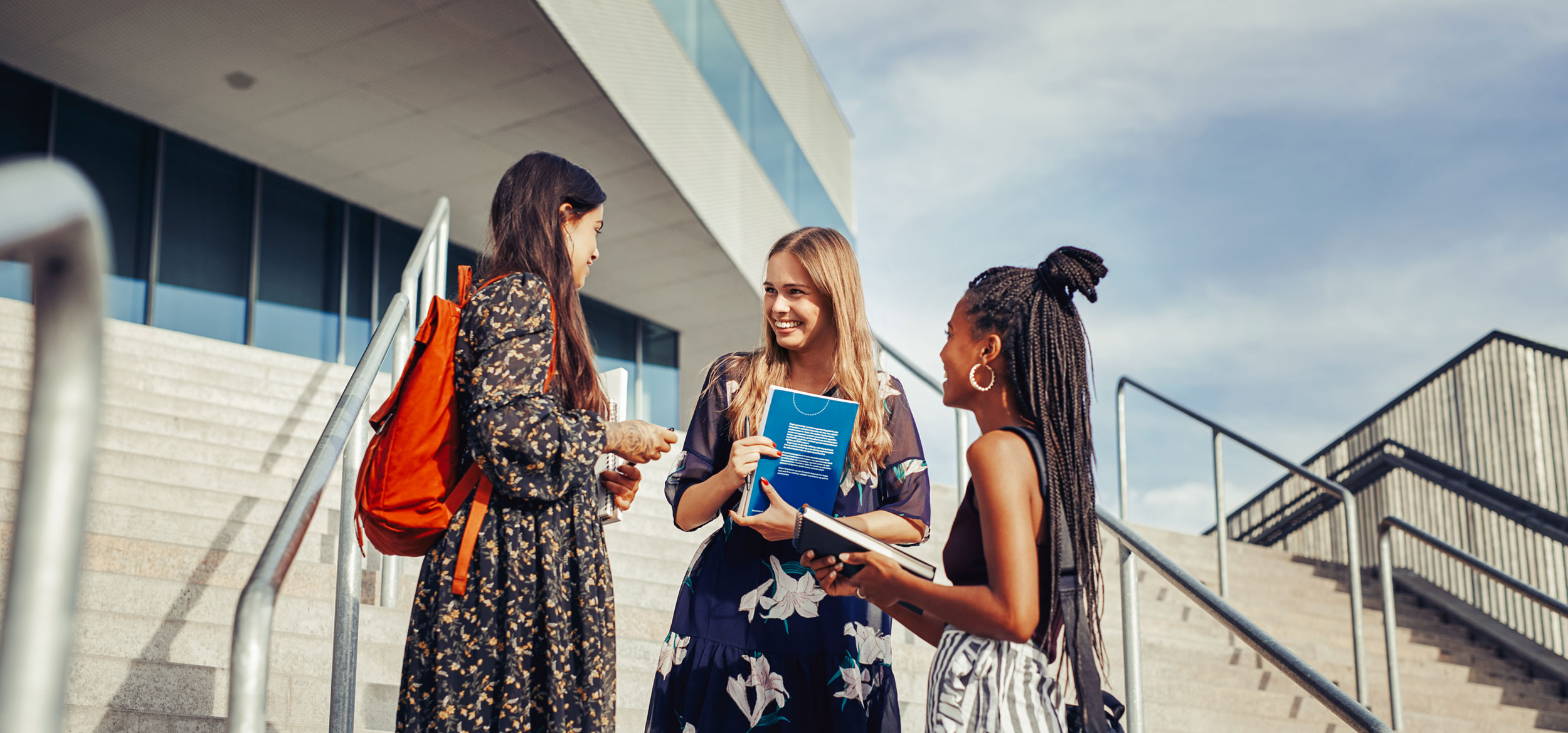 A group of students talking and standing on the steps of a campus. 