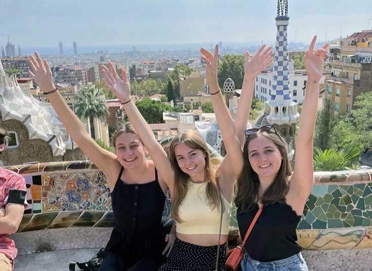 three students with arms raised sitting on the stone bench at Gaudi's Park Guell, Barcelona