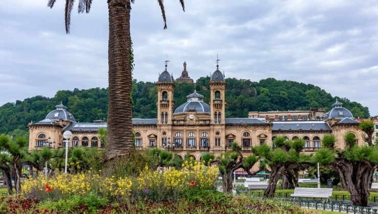 An ornate Spanish building behind a palm tree and lush garden on a cloudy day. 
