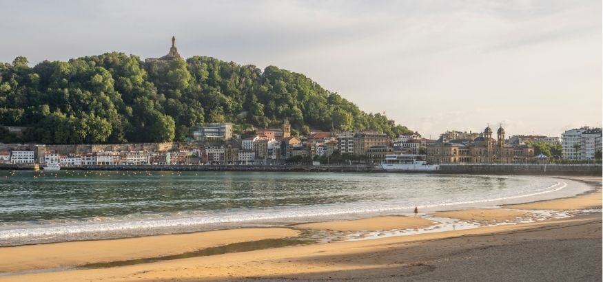 Old Spanish city buildings sit on a shoreline below a large tree-covered hill