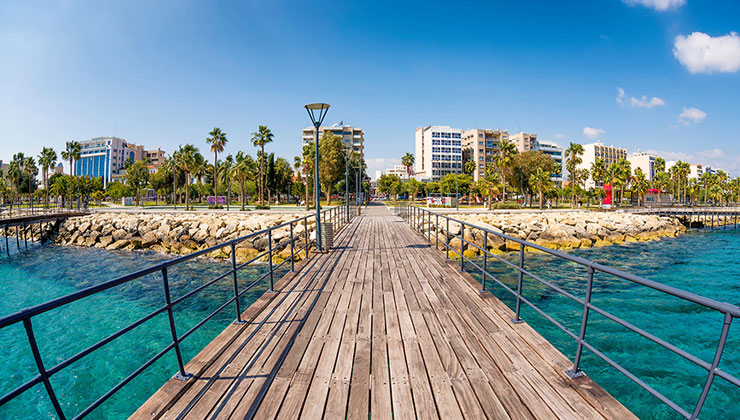 View of old wooden pier over aqua colored water with town and palm trees in the background at the Enaerios Seafront in Limassol, Cyprus.