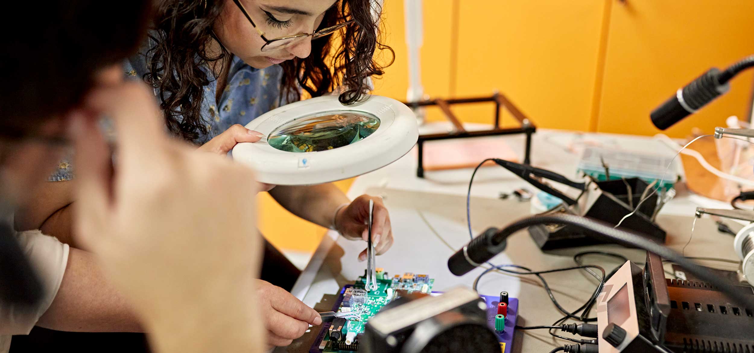 electrical engineering student looks through magnifying glass as they work on a circuit board.