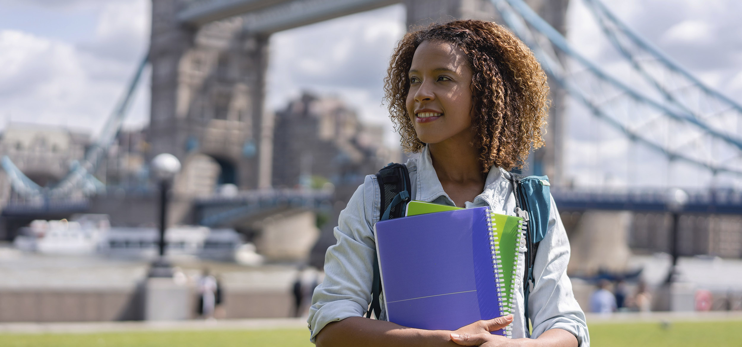 A student studying abroad in London smiles with their schoolwork in front of Tower Bridge. 