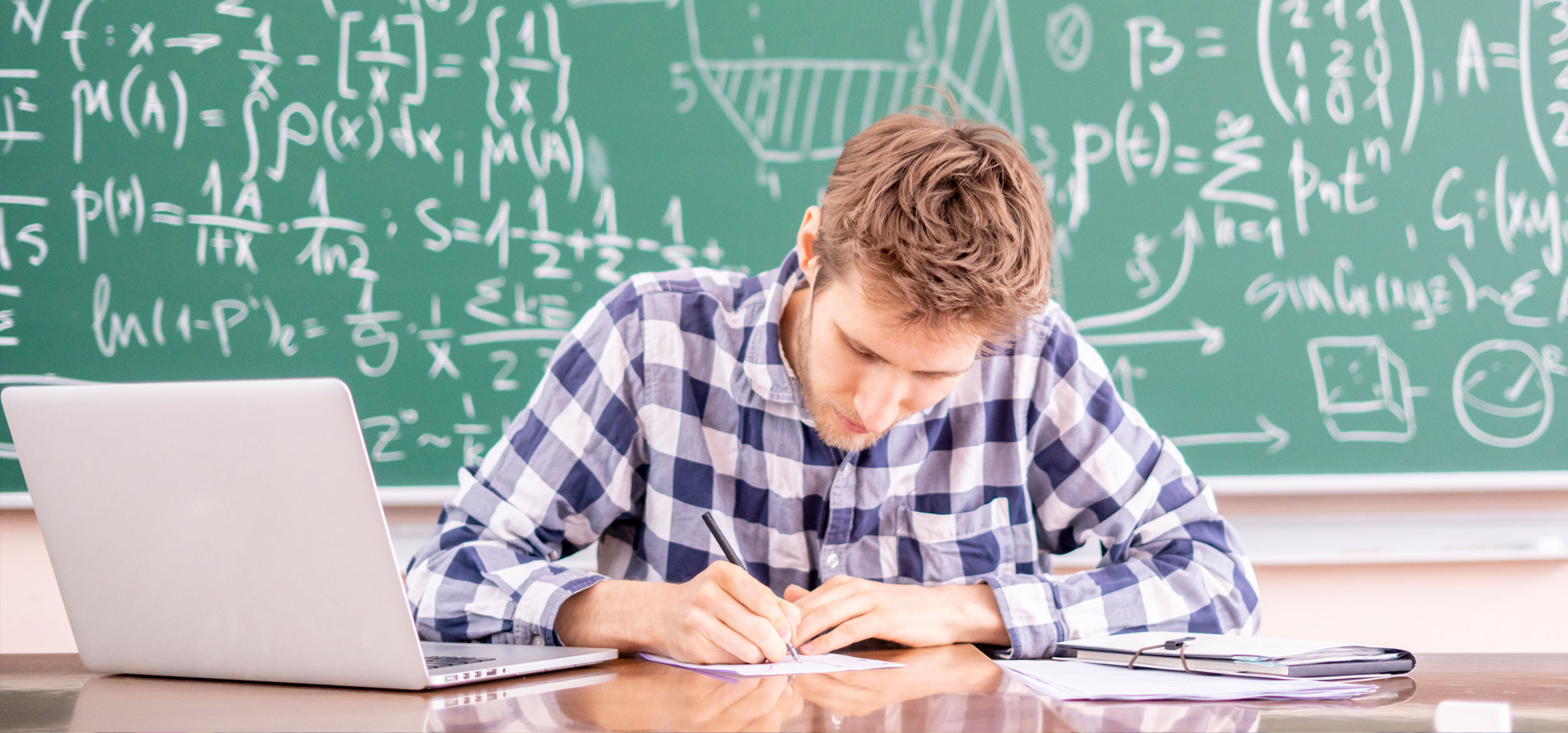 A student works out a math problem on paper while mathematical equations appear on a chalkboard behind the desk. 