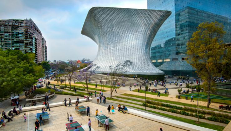 A large shiny metal building, shaped like an hourglass towers over a city center in Mexico City.