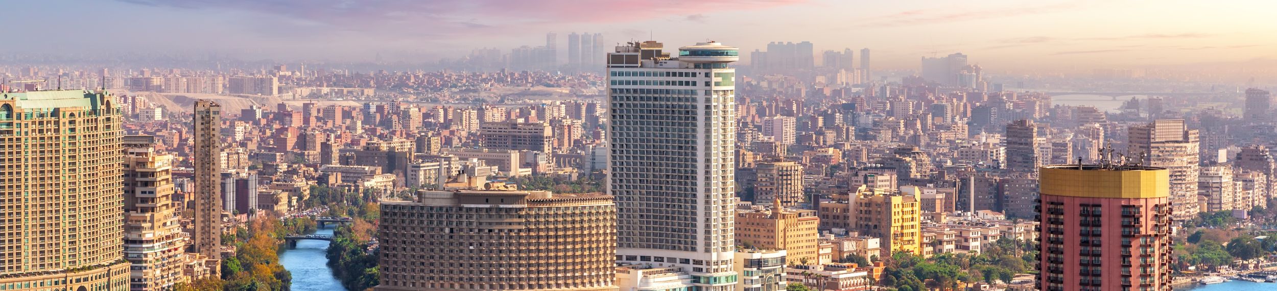 Aerial view of the skyline containing tall buildings over the Nile River, set before a pink and purple sky.