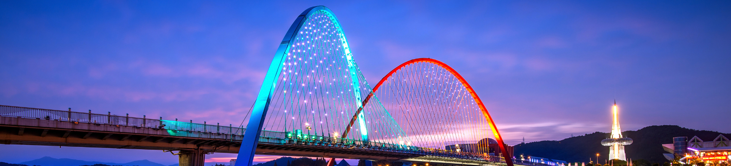 View of two overlapping red and blue-lit bridges at sunset, reflecting over water with city lights in the background.