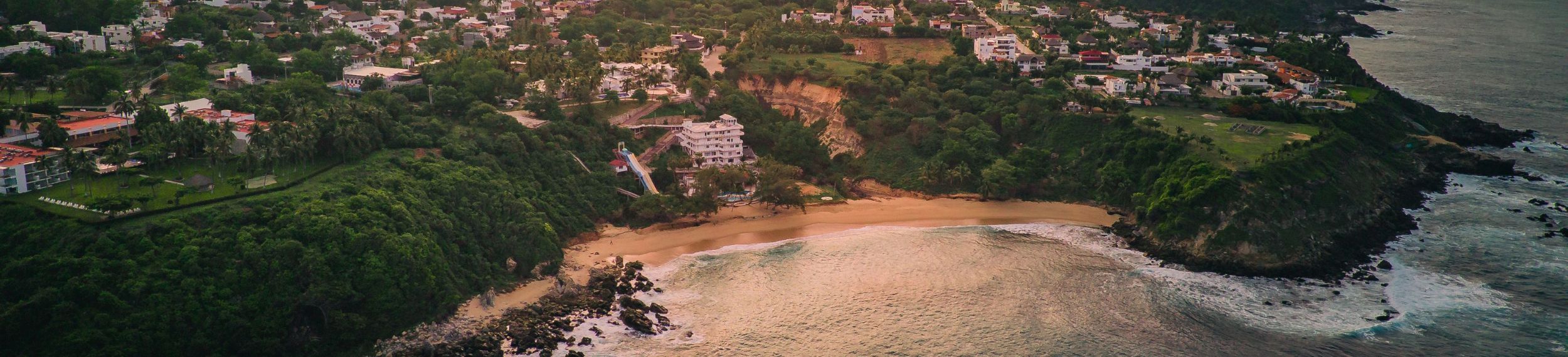 An aerial view of lush green trees and sand crashing into waves at Zicatela Beach in Puerto Escondido.