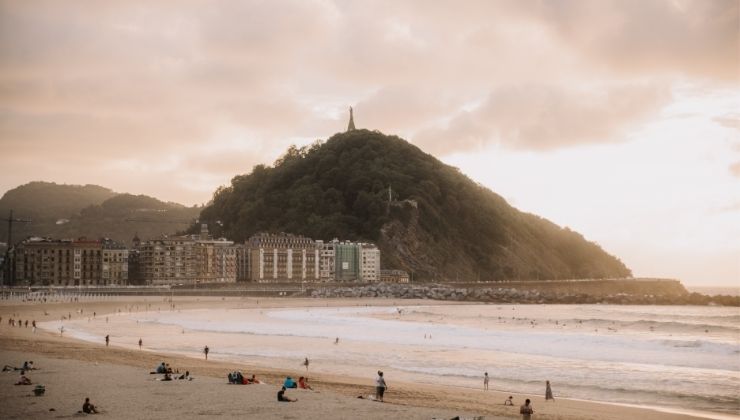 People relaxing on the sand and swimming in water at a beach during sunet, with a large grass-covered hill in the background. 