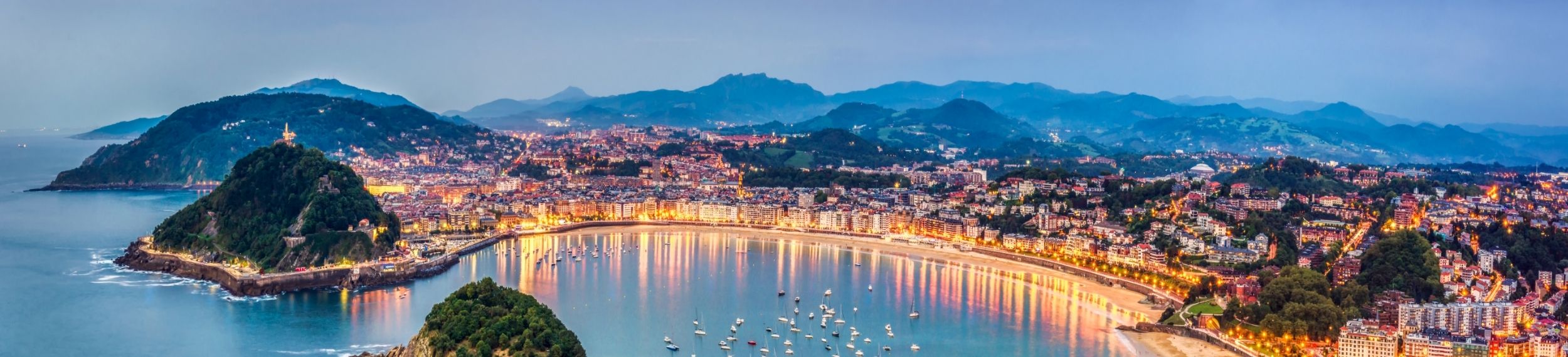 Panoramic view from above of a bay lit by city lights, with boats floating in the water