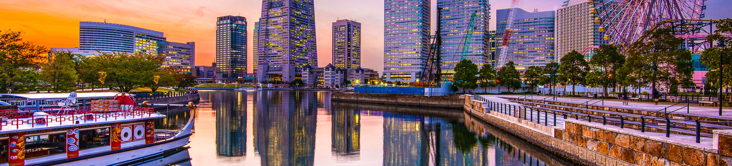 View of the bayside with a boat and ferris wheel in the foreground and city buildings in the background. 