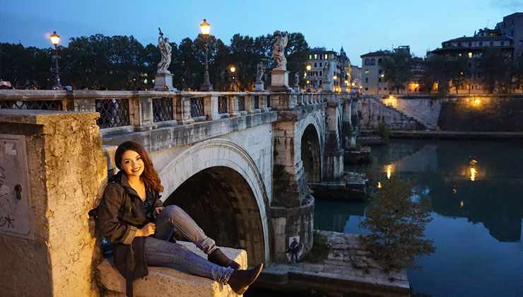 UC San Diego student on a ledge near a stone bridge a river at night in Rome, Italy.