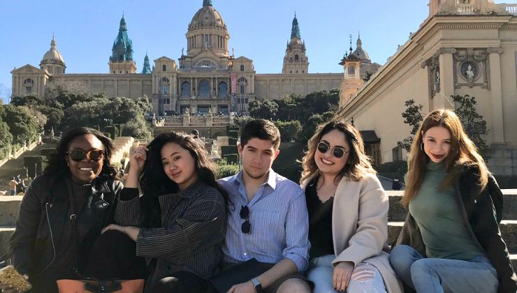 A group of students pose for a photo on a staircase with a large historic building in the background. 