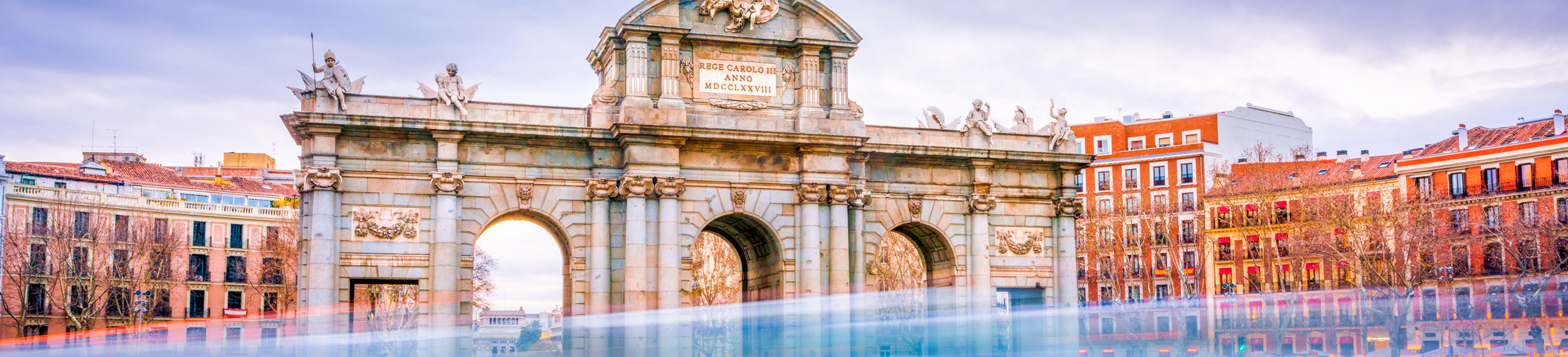 A shot of The Alcala Door (Puerta de Alcala), a post-Roman triumphal arch with cars passing by and buildings in the background in Madrid, Spain. 