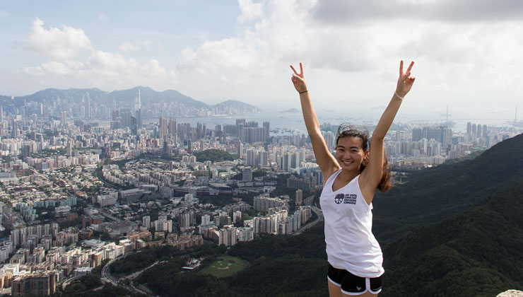 Student raises their hands in a double peace sign at Lion Rock with Hong Kong in the distance.