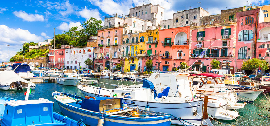 Blue and white boats sit in the aqua colored harbor with bright and colorful homes in Procida Island, Campania, Italy.