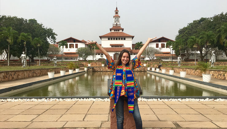 Student dressed in colorful attire, poses with her arms outstretched to the sky on the campus of the University of Ghana.