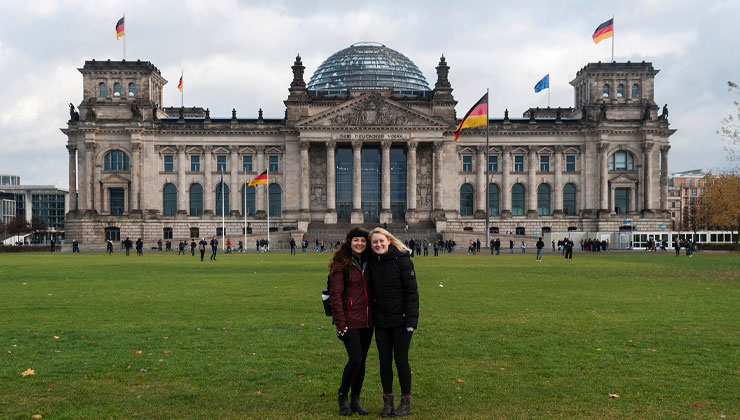 Two students stand on the grass on an overcast day in front of the Reichstag Building in Berlin, Germany.