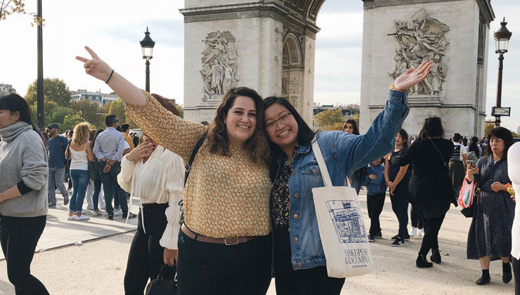 Two students posing with their arms out in front of the Arc de Triomphe in Paris, France 