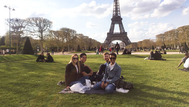 Four students sitting on grass in front of the Eiffel Tower. 