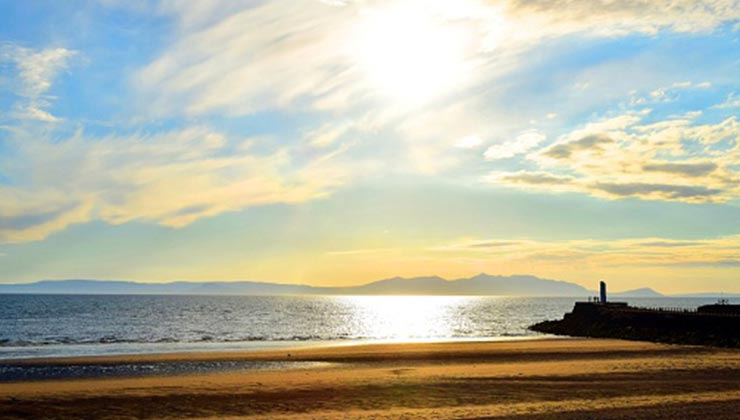 Ayr Beach in Scotland with silhouette of the harbor wall, shimmering sea, distant isle of Arran, the setting sun, and sky of pale yellow.