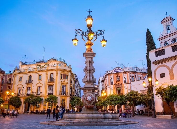People walk through the Plaza Virgen de los Reyes, behind a large lamppost at dusk in Seville