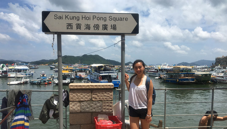 Student stands next to a sign by the water that reads Sai Kung Hoi Pong Square in Hong Kong.