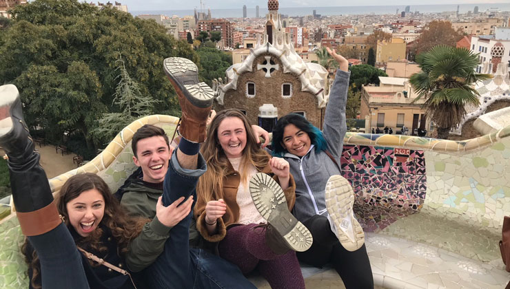 Four students sitting on the bench with their legs up in the air smiling at Park Güell in Barcelona, Spain.