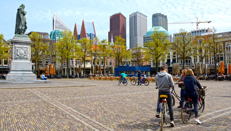 Het Plein square in spring, vieved from Binnenhof. In the foregruond pavement, on the right three cyclists. On the left statue of Willliam the Silent. Some modern skyscrapers in the very background. Horizontal image.