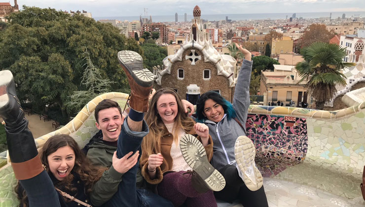 Four friends kick their feet up in the air and laugh at Park Güell in Barcelona, Spain.