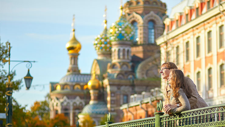 Young, smiling couple glow in the sunlight as they lean against a railing with the Church of Our Savior on the Spilled Blood behind them in St. Petersburg, Russia.