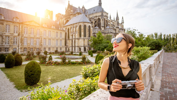 A female student holding a camera and looking to the side in front of Cathédrale Notre-Dame de Reims in Reims, France.