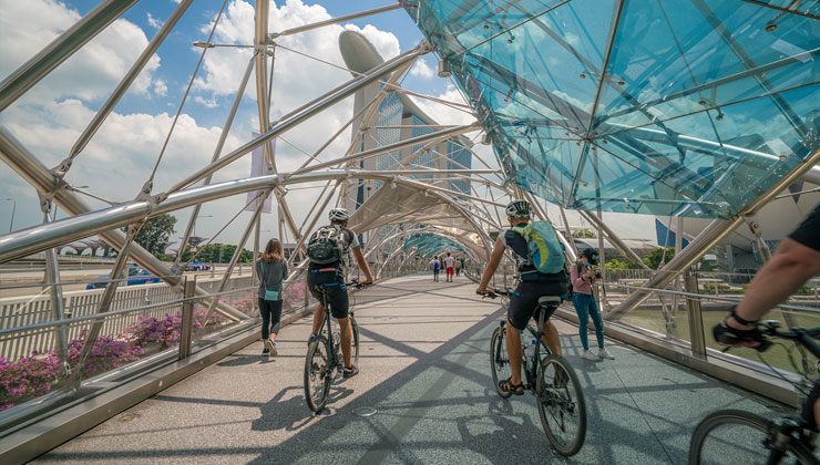 Bicyclists ride under the web of metal bars and blue glass of the Helix Bridge at Marina Bay in Singapore.