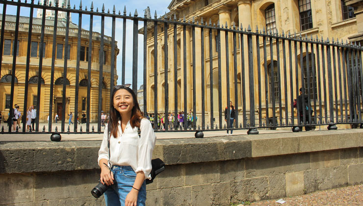 A student smiling on the University of Oxford campus near a fence and buildings in the background in Oxford, England. 
