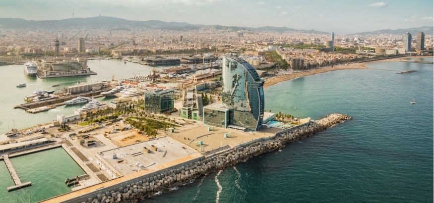 Aerial view of a half-circular skyscraper and other buildings on dock, shoreline and water in the background on a sunny day in Barcelona, Spain