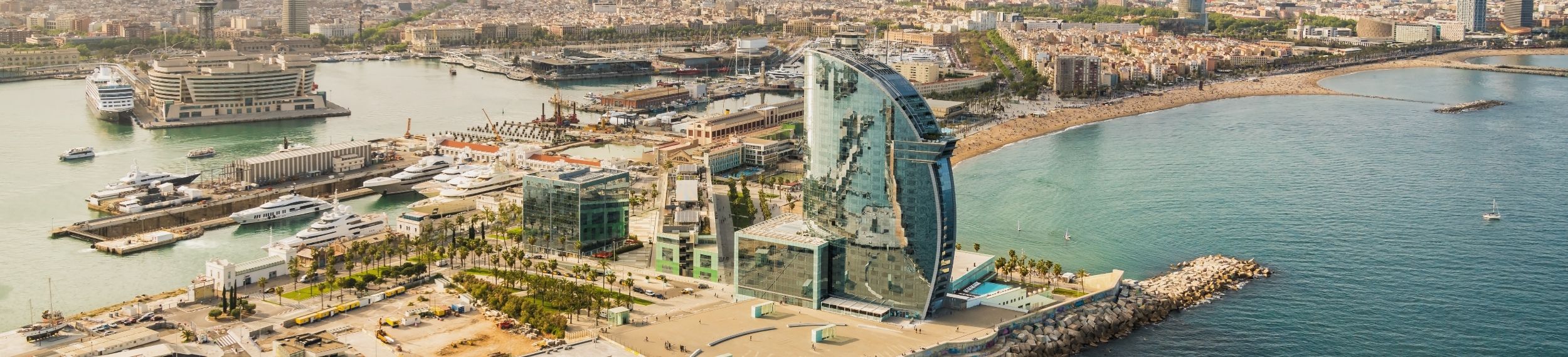 Aerial view of a half-circular skyscraper and other buildings on dock, shoreline and water in the background on a sunny day in Barcelona, Spain