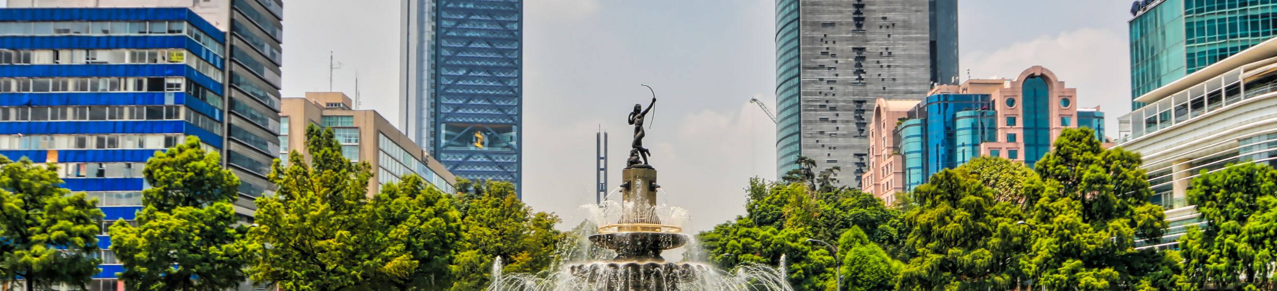 A statued fountain springs up in the middle of a city landscape with skyscrapers behind it.