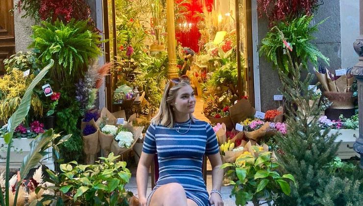 A person in a dress sits on a chair surrounded by plants and flowers in front of a colorful window.