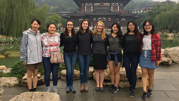 Eight students smiling at the camera with a pond and Chinese building in the background.  