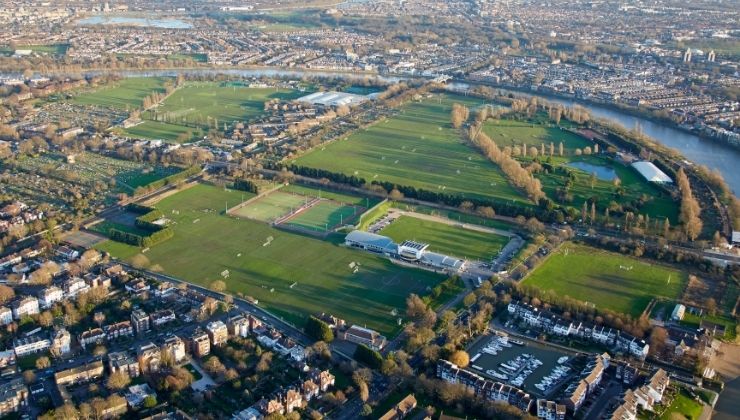 Aerial view of a large sports complex with multiple fields, surrounded by residential areas, roads, and a river in the background.