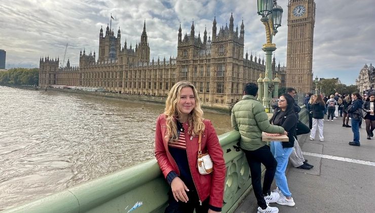 Person in red jacket leans on a railing and smiles for a photo in front of Big Ben clock tower and Westminster Palace. 