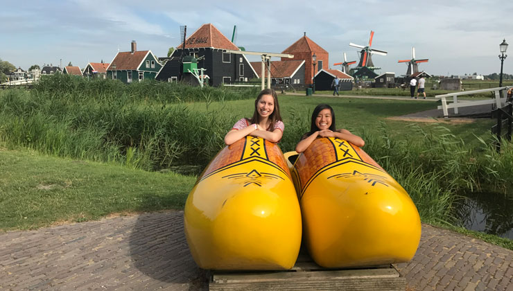 Two students sitting in giant clogs with a view of windmills and lush green grass at Wooden Shoe Workshop of Zaanse Schans in the Netherlands. 