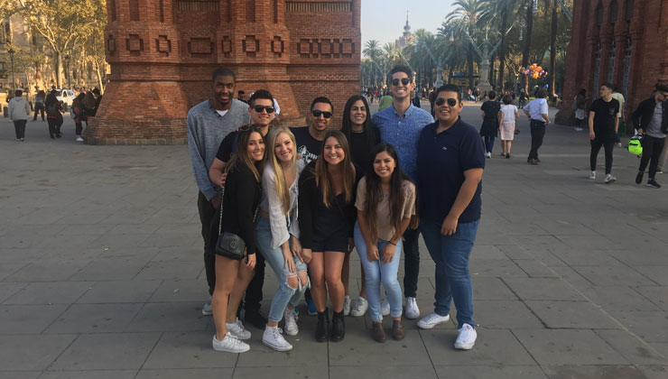 A group of ten students smiling at the camera with the view of the arch of the Arco de Triunfo de Barcelona in Barcelona, Spain.