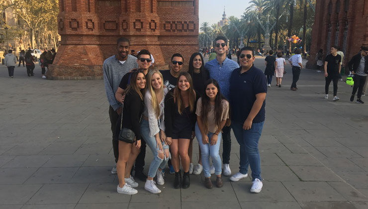 A group of ten students smiling at the camera with the view of the arch of the Arco de Triunfo de Barcelona in Barcelona, Spain. 